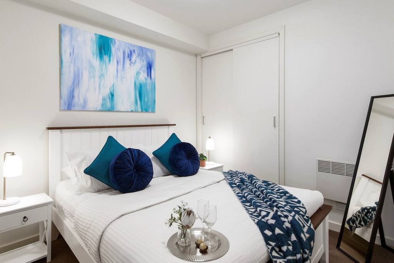 A hotel room with a bed, bedside tables, a nightstand, a bedside lamp, a wall-mounted abstract painting, a wardrobe, and a mirror.