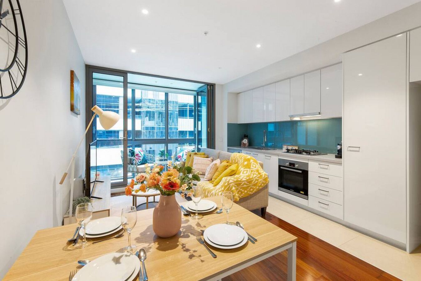 A modern kitchen with white cabinets, a dining area with a wooden table set for four, and a large window offering a view of the cityscape.