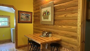 Eine Holzwand mit zwei gerahmten Bildern, ein Holztisch mit zwei Hocker und ein Fenster mit Blick auf Grünflächen.