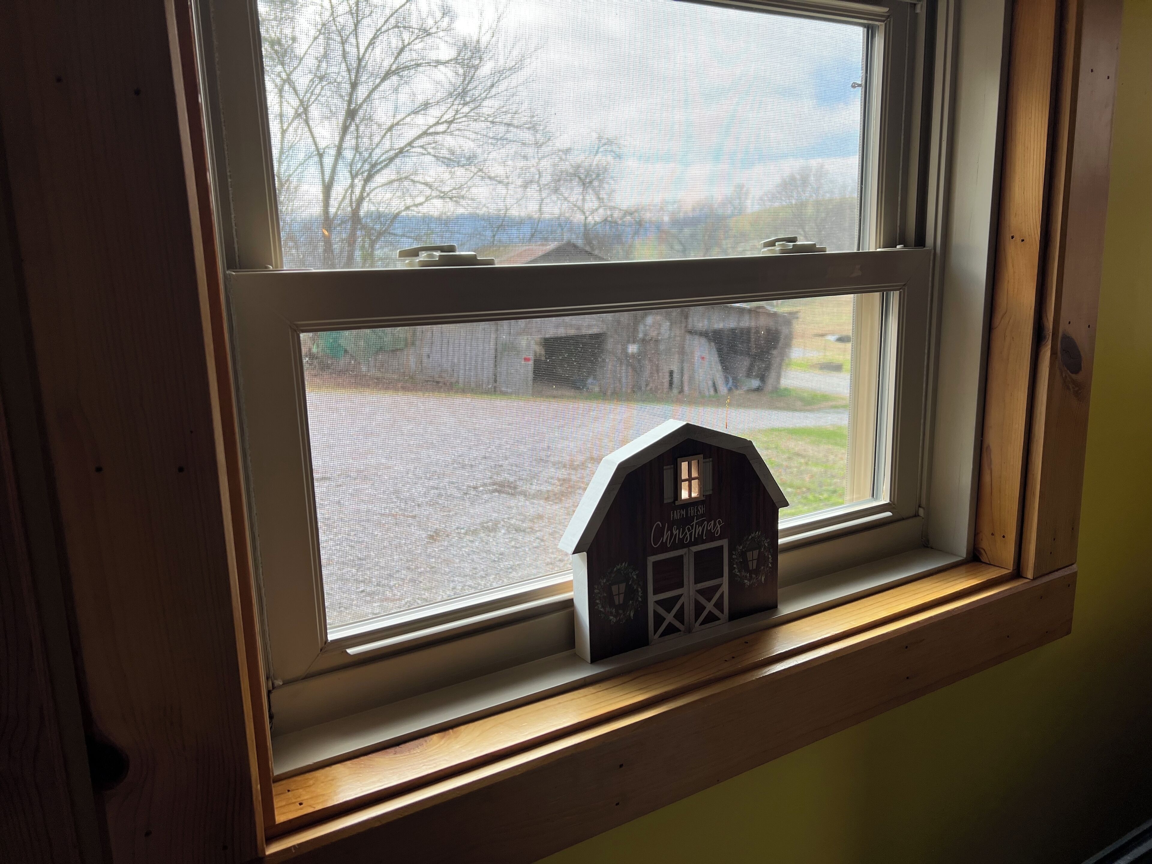 A window with a view of a rural landscape, a small wooden house model labeled "Farm Fresh Christmas," and a wooden window frame.