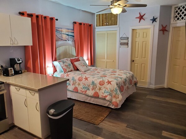 A bedroom with a bed, a desk with a coffee maker, a trash bin, and a ceiling fan.