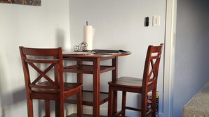 Un coin repas avec une table ronde et quatre chaises en bois. Un tableau est accroché au mur, ainsi que deux assiettes décoratives. Un appareil de climatisation est installé au-dessus de la table.