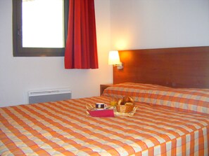 Une chambre d’hôtel avec deux lits, une fenêtre avec des rideaux rouges, une lampe fixée au mur et un panier contenant des articles pour le petit-déjeuner, posé sur l’un des lits.