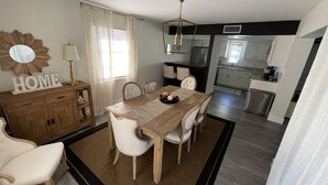 Ein Essbereich mit einem Holztisch, weißen Stühlen, einer Pendelleuchte und einer Küche im Hintergrund.