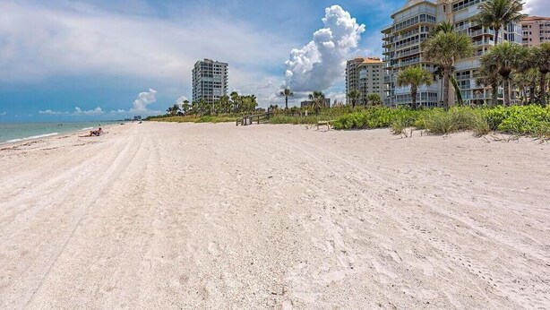 Ein weiter Sandstrand, bewölkter Himmel und hohe Gebäude mit Balkonen im Hintergrund, sowie einige Palmen.
