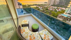 A balcony with a wicker chair and cushions, overlooking a cityscape and a body of water during sunset. - Exclusive Apartment 3 Bedroom in Beach Club (Santa Marta)