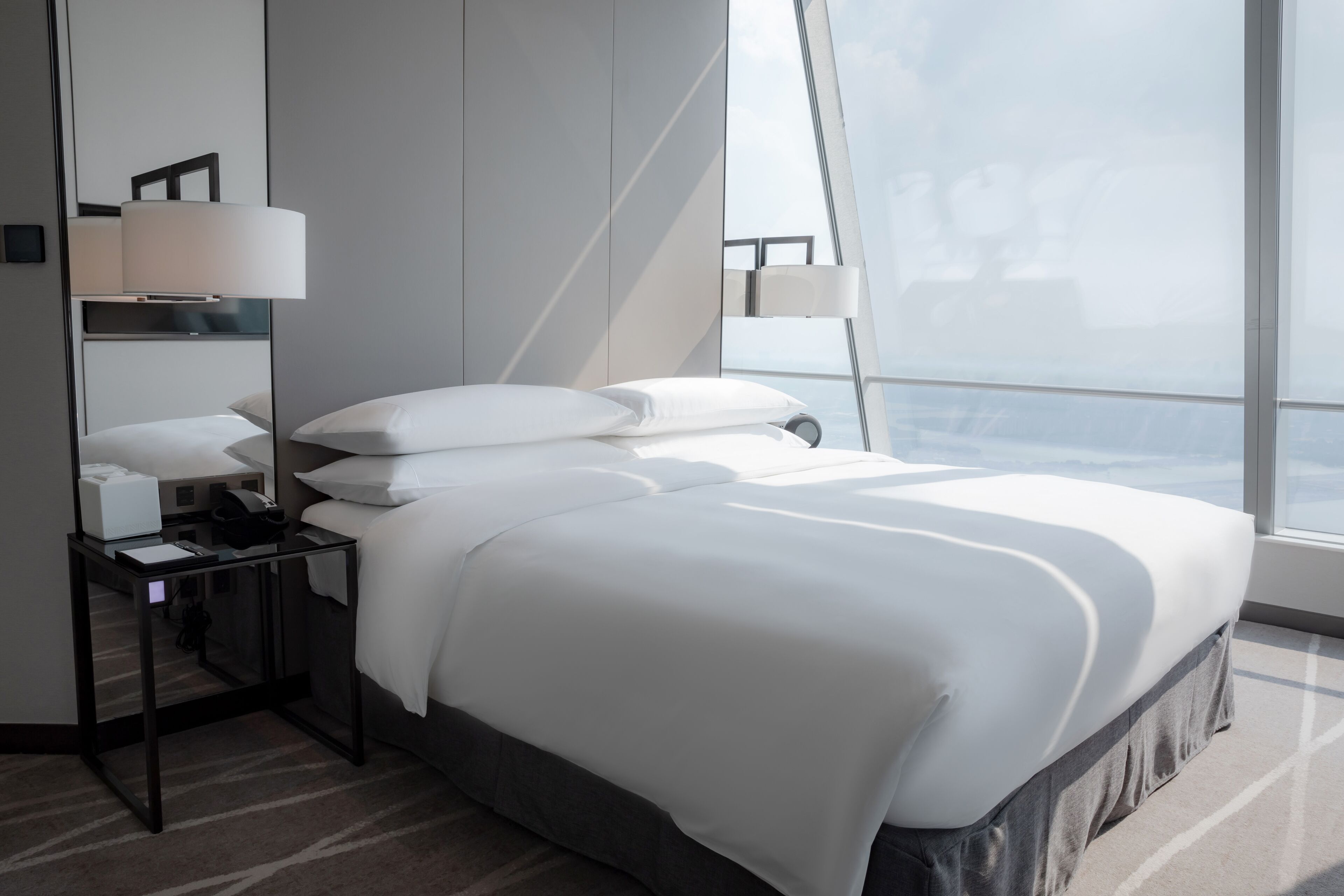 Habitación de hotel moderna con una cama grande, una mesita de noche con lámpara, un espejo y vistas a la ciudad.