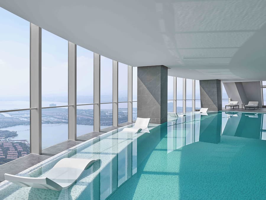 A modern indoor swimming pool with clear glass walls, white lounge chairs, and a view of a cityscape and water body.