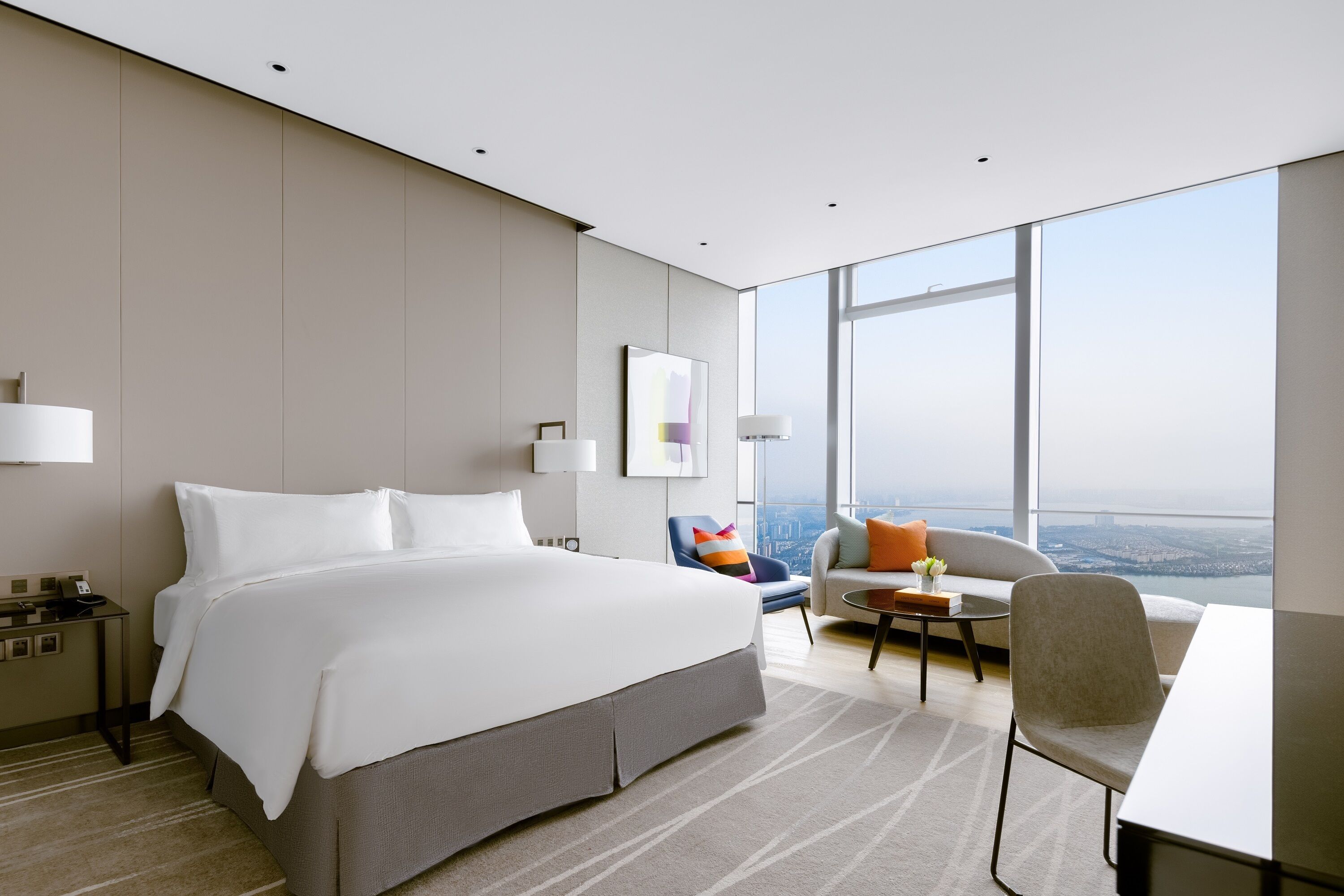 A modern hotel room with a large bed, a desk, a chair, a sofa, and a view of the city.