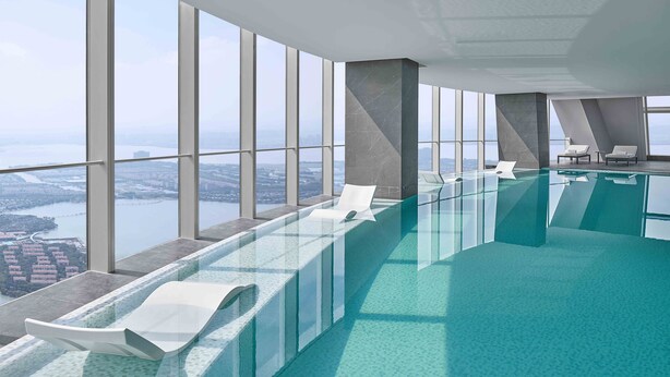 Una piscina interior moderna con paredes de vidrio transparente, sillas reclinables blancas y vistas a la ciudad.
