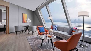 Una habitación de hotel moderna con un amplio ventanal panorámico, un sofá, dos sillones, una mesita con una botella y flores, y una lámpara de pie.