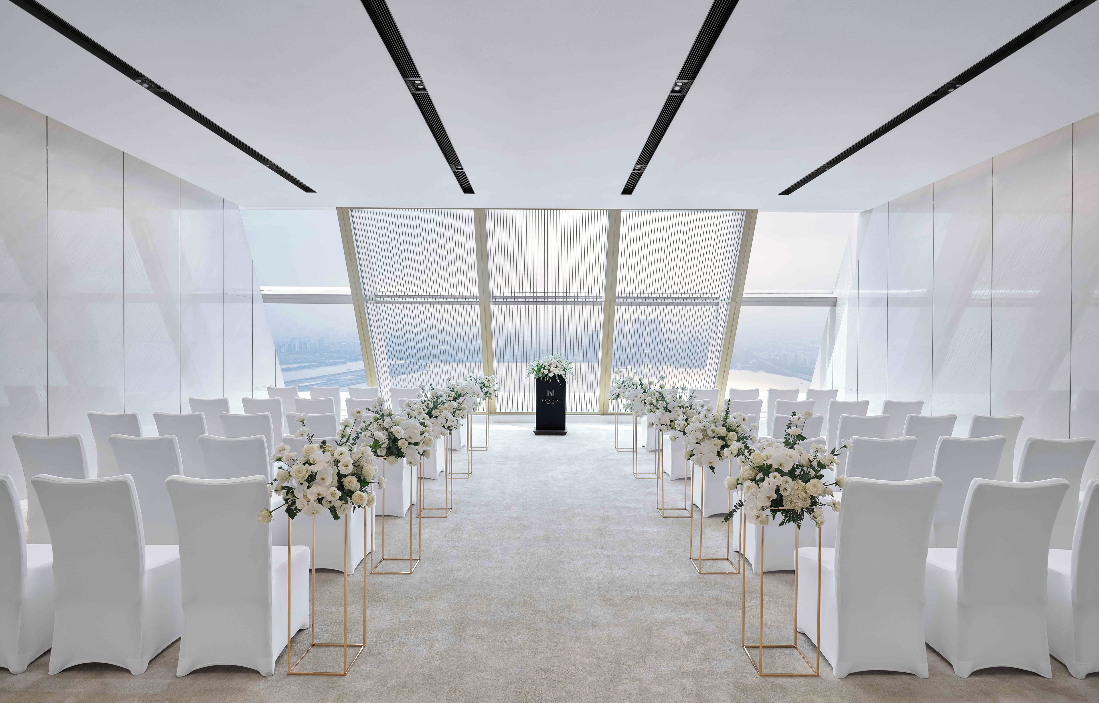 A modern indoor venue with white chairs arranged in rows, a podium at the front, and a view of the cityscape through large windows.