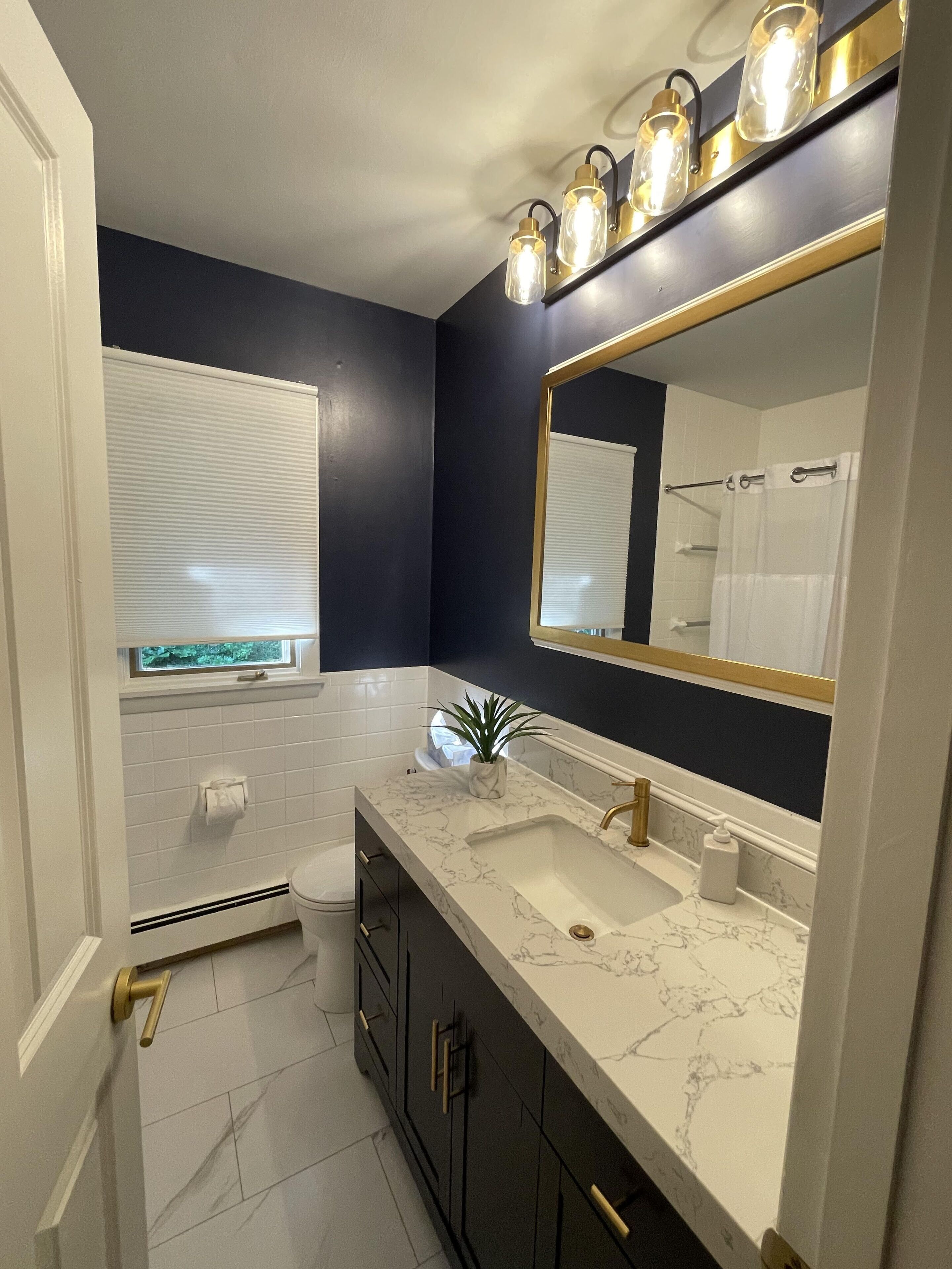 A bathroom with a dark blue wall, white tiled floor, a white toilet, a sink with a marble countertop, and a mirror with a gold frame.