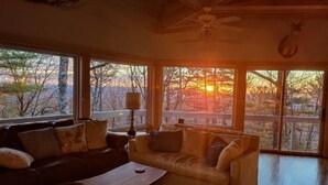 A cozy living room with a sofa and a coffee table, large windows with a view of trees and a sunset, and a ceiling fan.