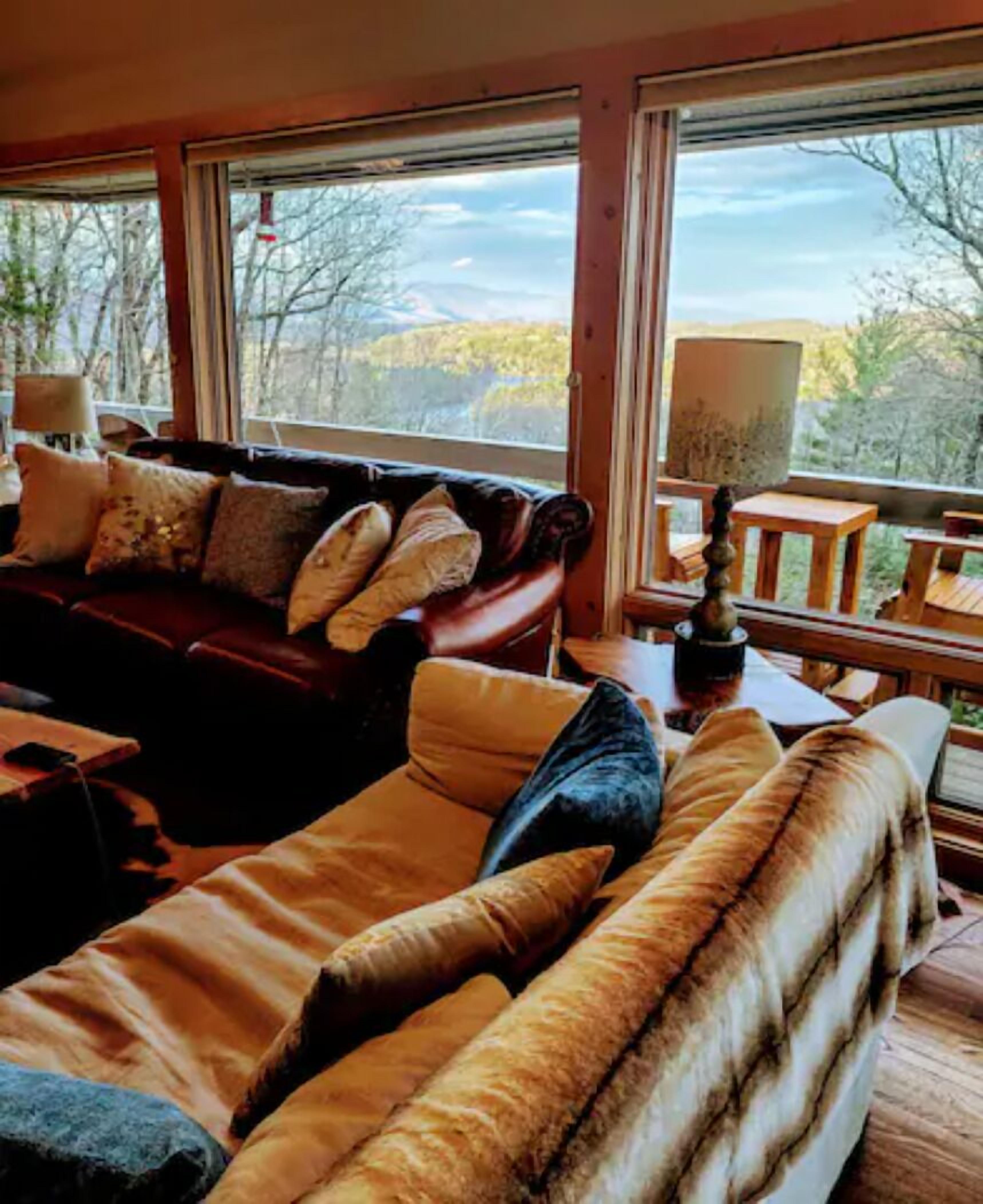 A cozy living room with a leather sofa and a fur throw, large windows with a view of trees and mountains, and a wooden table with a lamp.