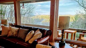 A cozy living room with a leather sofa and a fur throw, large windows with a view of trees and mountains, and a wooden table with a lamp.
