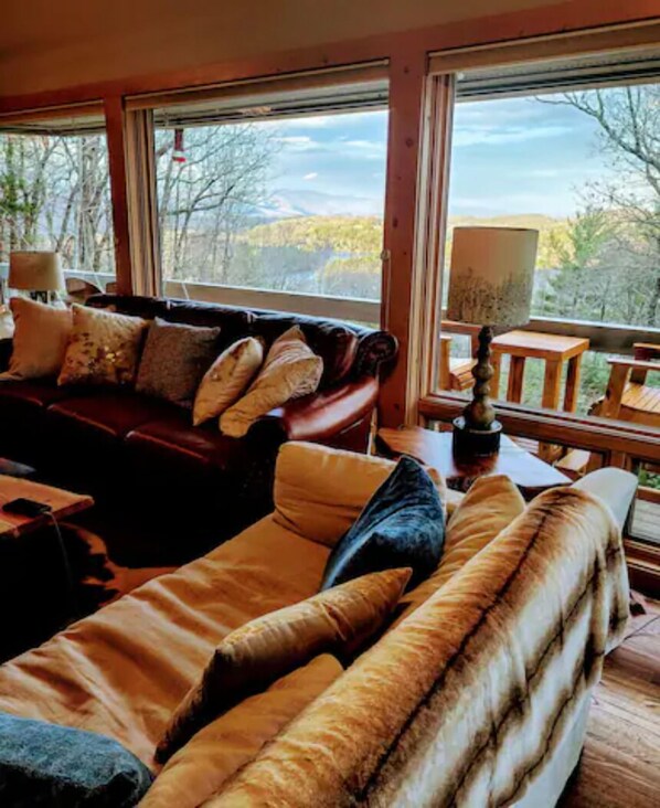 A cozy living room with a leather sofa and a fur throw, large windows with a view of trees and mountains, and a wooden table with a lamp.
