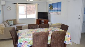 Un salon avec un canapé, une table à manger entourée de chaises en osier, un téléviseur posé sur un meuble en bois et un tableau sur le mur représentant un paysage de plage.