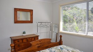 Une chambre avec une commode en bois, un lit recouvert d’une couvre-lit à motifs floraux, une tête de lit en bois, un miroir, un étendoir à linge et une fenêtre avec des stores.