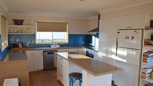 A modern kitchen with white cabinets, a stainless steel dishwasher, a built-in oven, and a refrigerator. There is a window above the sink and a stool near the counter. - Amberlee House - South West Rocks (SOUTH WEST ROCKS)