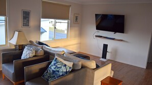 A modern living room with a sofa set, a television mounted on the wall, and a window with blinds. - Amberlee House - South West Rocks (SOUTH WEST ROCKS)