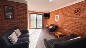 A living room with brick walls, a television mounted above a fireplace, and a sliding glass door leading to an outdoor area. - Cameo Court Unit 7  - South West Rocks (SOUTH WEST ROCKS)