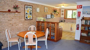 A cozy kitchen with a brick wall, wooden cabinets, a dining table with chairs, a microwave, and a bookshelf. - Sunny Nook - South West Rocks - Bring your own linen (SOUTH WEST ROCKS)