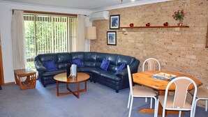 A living room with a black leather sofa, a wooden coffee table, and a dining table with chairs. There is a brick wall with a shelf holding decorative items. - Sunny Nook - South West Rocks - Bring your own linen (SOUTH WEST ROCKS)