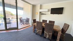 A room with a dining table and chairs, a flat-screen TV, and a sliding glass door leading to an outdoor area. - Ocean View 1 - South West Rocks - Bring your own linen (SOUTH WEST ROCKS)