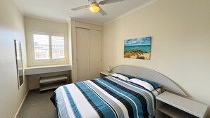A bedroom with a bed, a desk, a ceiling fan, a window with blinds, and a painting of a beach scene. - Ocean View 1 - South West Rocks - Bring your own linen (SOUTH WEST ROCKS)