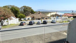 A coastal road with parked cars, houses, and a view of the sea and mountains in the distance.