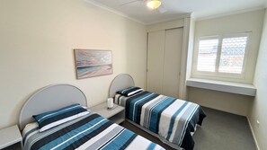 A bedroom with two single beds, a bedside table with a lamp, a window with blinds, and a painting on the wall.