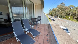 A balcony with sun loungers, a small table, and chairs, overlooking a road with parked cars and trees. - Ocean View 1 - South West Rocks - Bring your own linen (SOUTH WEST ROCKS)