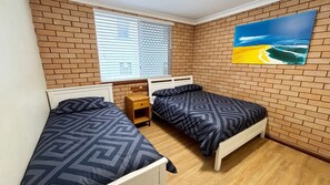 Een kamer met twee bedden, een raam met zonwering, een houten nachtkastje en een schilderij aan de muur.