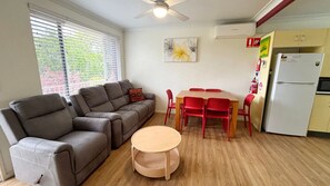 Een knus woongedeelte met een bank, een klein rond tafeltje, een eettafel met stoelen, een plafondventilator en een koelkast.