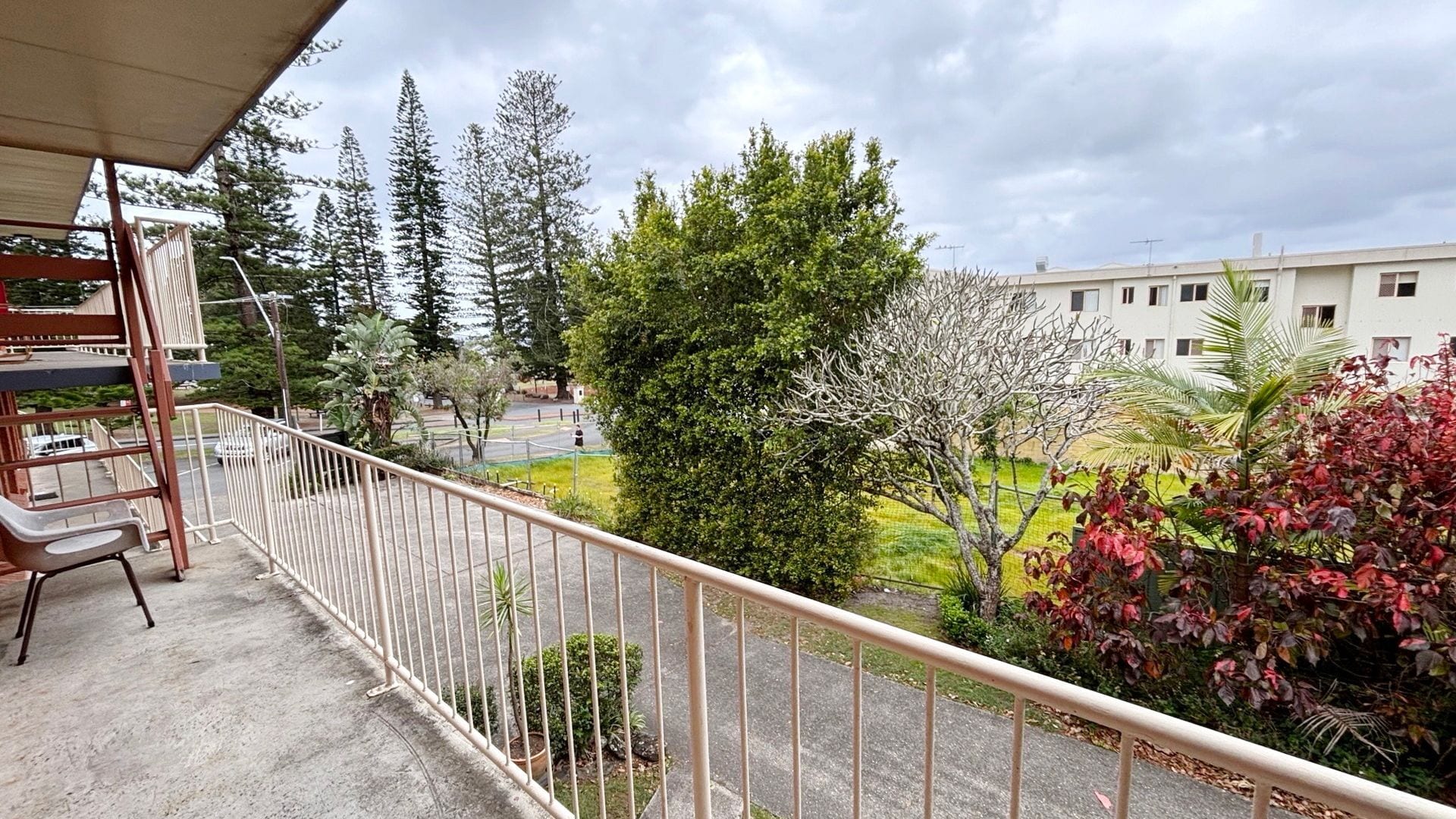 A balcony with a metal railing overlooking a courtyard with trees and a building in the background.