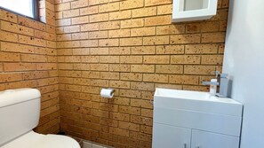 A bathroom with a brick wall, a toilet, a sink, and a mirror. - Seacrest # 5  - South West Rocks - Bring your own linen (SOUTH WEST ROCKS)