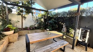 An outdoor patio with a wooden table and bench, surrounded by potted plants and a barbecue grill. - Landwin Gardens 1 - walk to the beach (SOUTH WEST ROCKS)