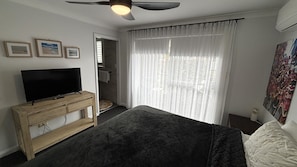 A bedroom with a bed, a TV on a wooden stand, a ceiling fan, a window with sheer curtains, and a painting on the wall.