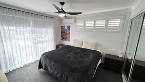 A bedroom with a bed, a ceiling fan, a window with blinds, a painting on the wall, and a door with a mirror. - Landwin Gardens 1 - walk to the beach (SOUTH WEST ROCKS)