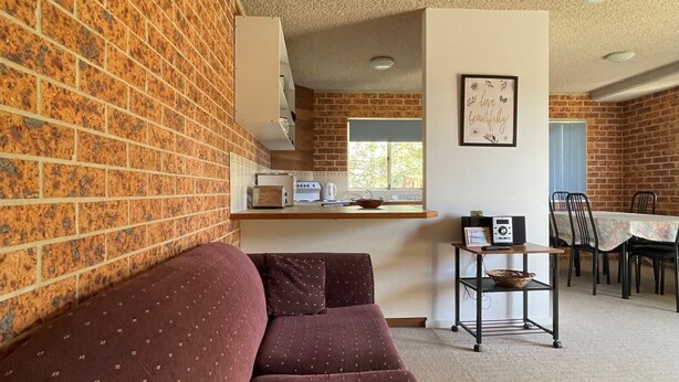 A cozy living area with a brick wall, a sofa, a small kitchen with a toaster and kettle, a dining table with chairs, and a framed sign on the wall.