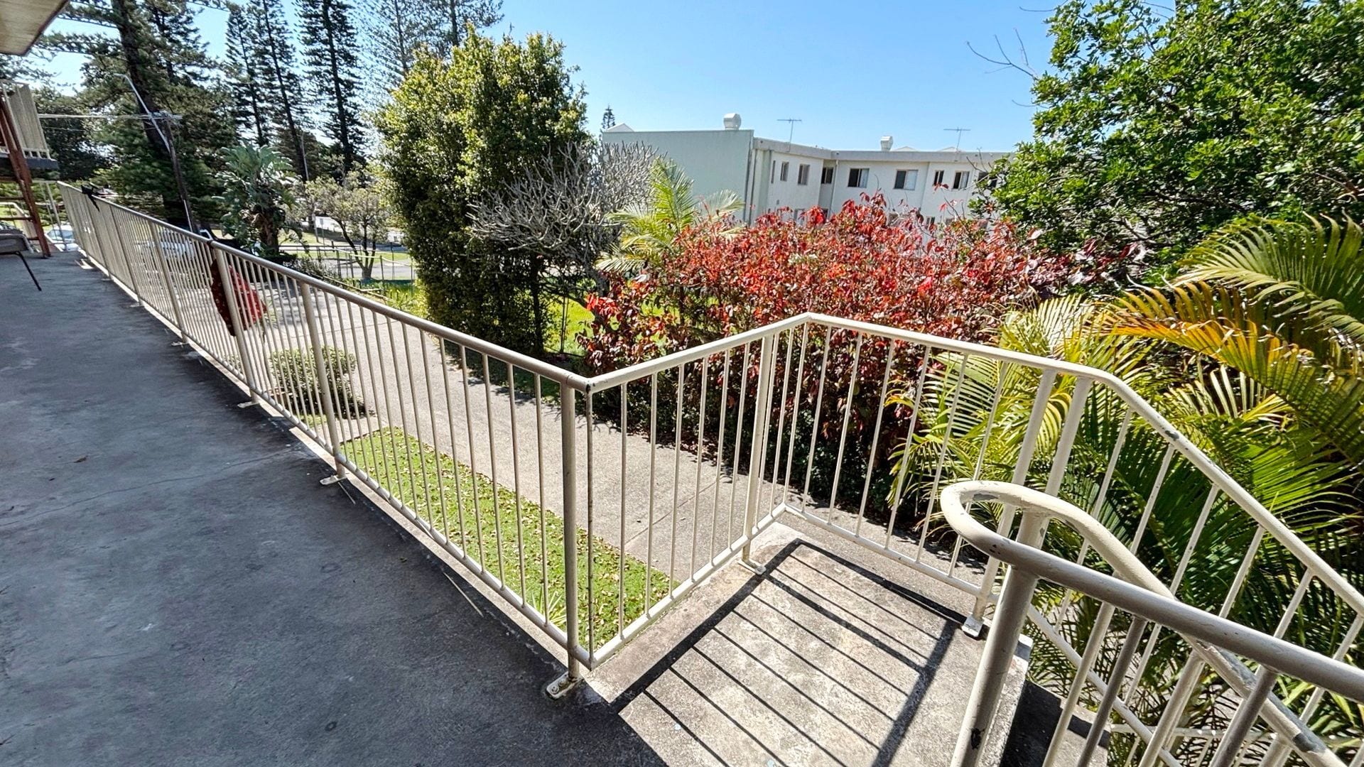 A balcony with a metal railing, a concrete floor, and a view of a building and trees.