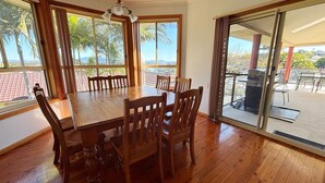 A dining area with a wooden table and chairs, large windows with curtains, and a view of palm trees and mountains. - Horizons - South West Rocks - Bring your own linen (SOUTH WEST ROCKS)