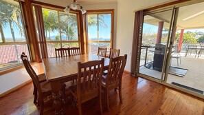 A dining area with a wooden table and chairs, large windows with curtains, and a view of palm trees and mountains. - Horizons - South West Rocks (SOUTH WEST ROCKS)
