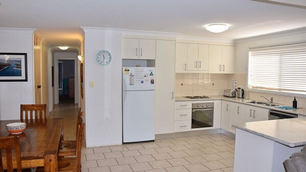 A compact kitchen with white cabinets, a built-in oven, a sink, and a dining area with a wooden table and chairs. - Parkside # 1 - South West Rocks (South West Rocks)
