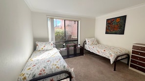 A bedroom with two single beds, a dresser, a painting, and a window with a view of buildings. - Parkside # 1 - South West Rocks (South West Rocks)