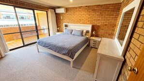 A bedroom with a bed, a bedside table, a dresser, and a large window with a view of a building.
