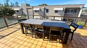Uma área de refeições ao ar livre coberta, com mesa e cadeiras, guarda-corpo de vidro e um edifício ao fundo.