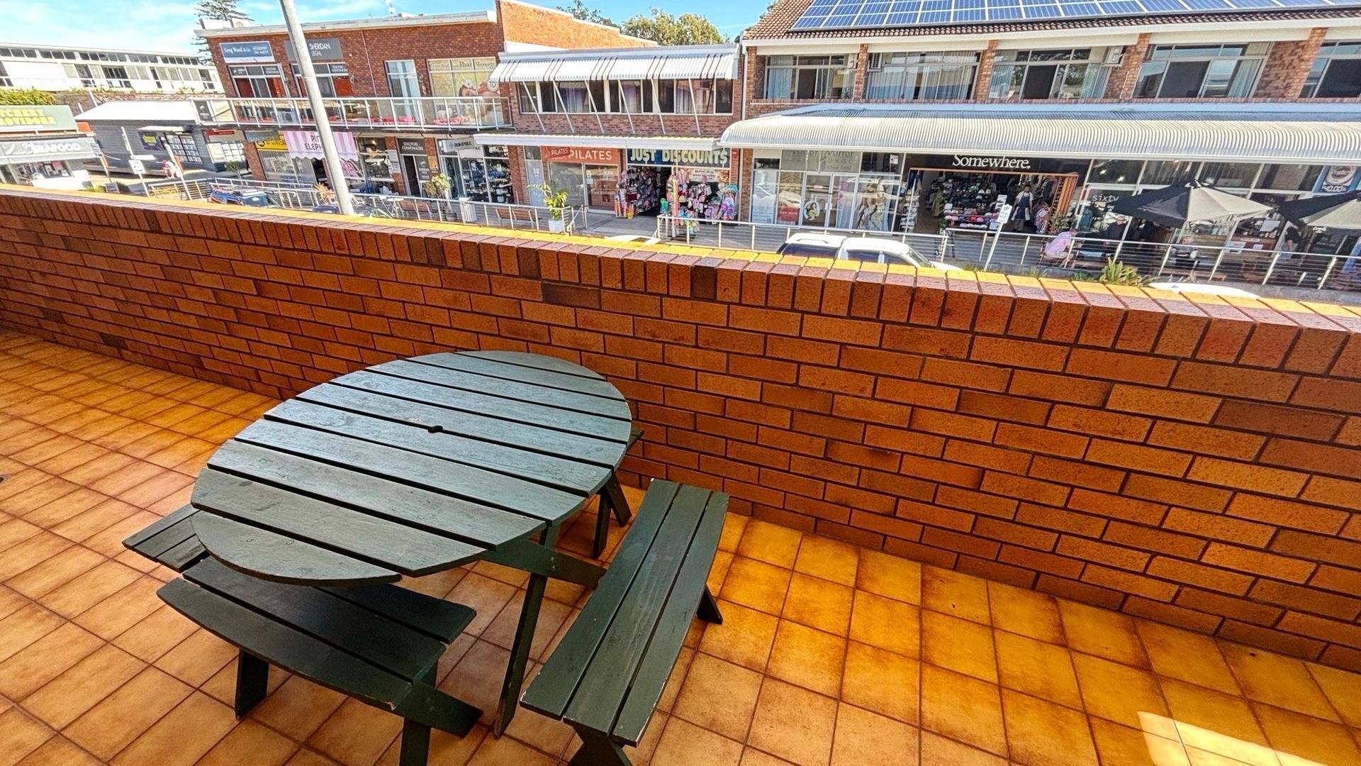 A brick wall with a wooden table and benches, overlooking a street with shops and a building with solar panels.