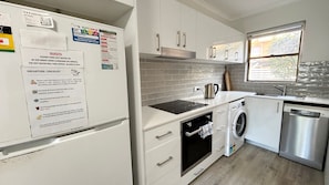 Una cocina moderna con armarios blancos, horno empotrado, lavadora y un lavabo. Hay varios avisos y una lista de salida en el refrigerador.